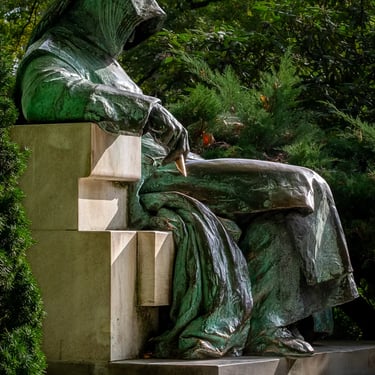 Side view of the hooded Anonymous statue under leafy trees, seated in thought.