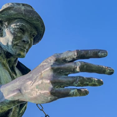 Imre Soós statue with hand and head extended forward, lit sharply beneath blue sky.