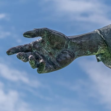 Reverse view of outstretched bronze hand, silhouetted against clouds and open sky.
