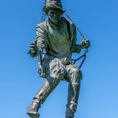 Full Imre Soós statue against sweeping blue sky and clouds, body leaning into motion.