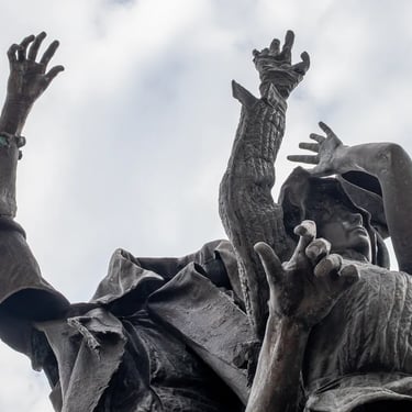Upward view of three bronze figures leaping into emptiness, hands flung up in loss.
