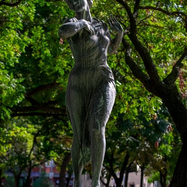 Full bronze statue of a young woman symbolizing spring, set among city buildings.