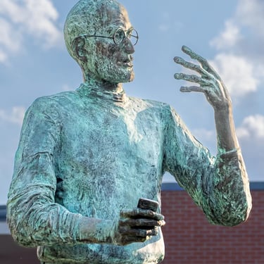 Steve Jobs statue showing upper body and both hands, with a cloudy blue sky background.