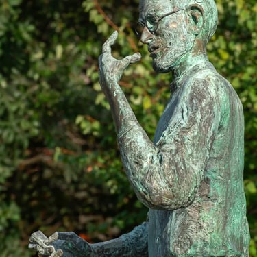 Side view of upper body of Steve Jobs statue, framed by green trees in the background.