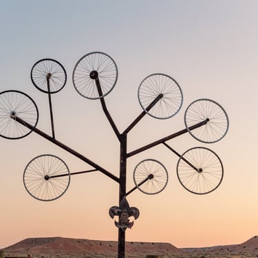 A metal sculpture shaped like a tree with bicycle wheels as branches against a desert sunrise sky.