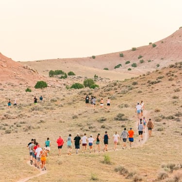 A group of diverse people trail running on a winding dirt path through desert hills at sunrise.