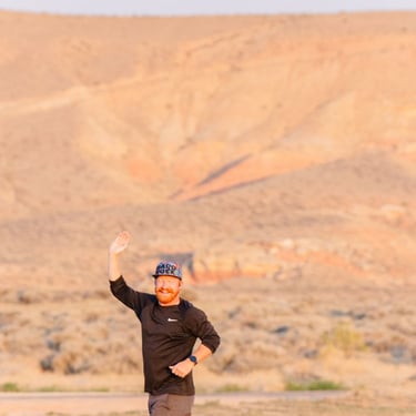 Smiling man with red beard trail running through a desert landscape at sunrise.