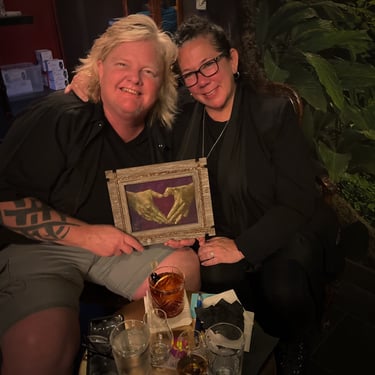 Two people smiling together, holding a framed artwork of golden hands forming a heart.