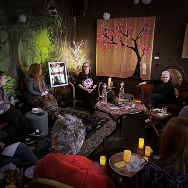 A group of people gathered in a cozy, dimly lit room with candles, artwork, and nature-themed decor, engaged in a discussion.