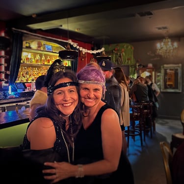 Two women in 1920s-themed outfits smiling and embracing at a vintage-style bar. with chandeliers, mirrors, and a liquor shelf