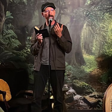 A man in a hat and glasses stands on a stage in front of a forest backdrop, holding a microphone and performing.