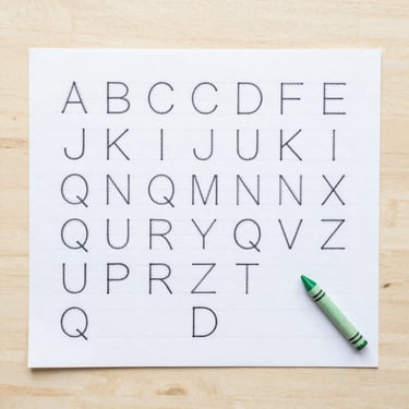 Overhead flat-lay of a printed alphabet tracing worksheet on a light wood table, completed with green crayon, bright natural daylight