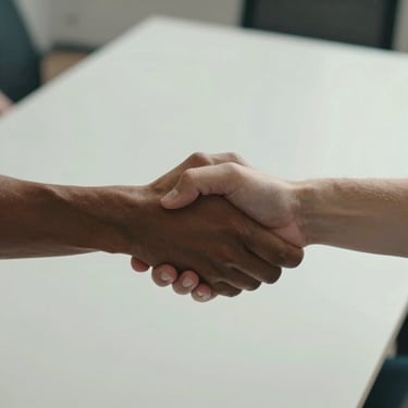 Two hands meeting in a firm handshake, one from a person with darker skin and one from a person with lighter skin, photographed from above at a studio desk with natural daylight. Clean minimalist workspace visible in soft focus behind. Emphasis on the gesture of trust and partnership.