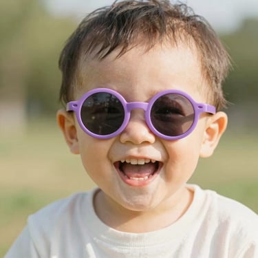 Close-up of a toddler laughing outdoors, wearing cute round purple sunglasses, bright warm sunlight, happy expression