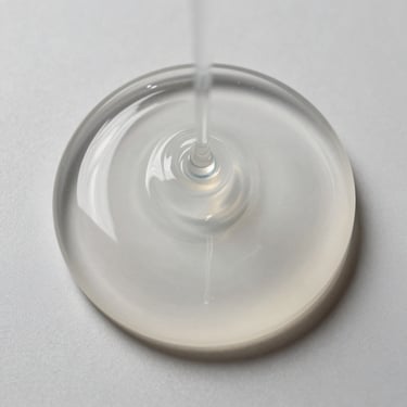 Top-down macro photography of pure, clear silicone fluid showing high viscosity and clean light refraction on a neutral clinical background, sharp shadows