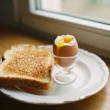 Soft boiled eggs in a small glass cup next to toasted buttered bread on a vintage plate, warm morning light, nostalgic breakfast scene