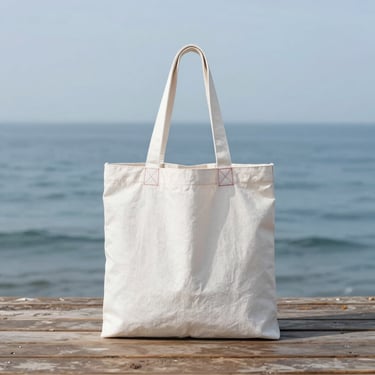 A minimalist white sailcloth tote bag with red stitching details, placed on a weathered wooden deck with soft ocean background