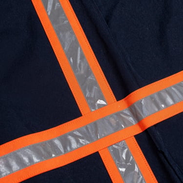 Close-up of high-visibility reflective strips and orange double-stitching on a dark navy PEMEX uniform fabric, industrial workshop lighting