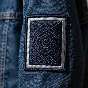 Macro close-up of a dense embroidered patch with a thick merrowed border stitched onto a heavy denim jacket sleeve, dramatic side lighting, shallow depth of field