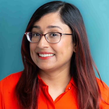 A woman with long, reddish-brown hair and glasses smiles warmly in a bright orange top against a teal background.