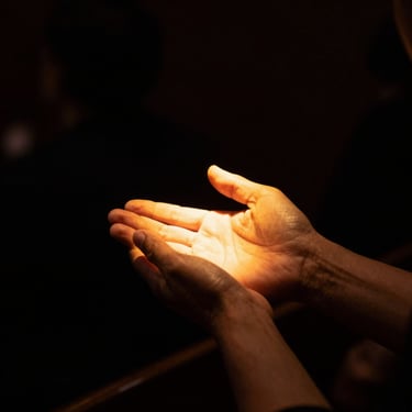 A hand holding another hand gently under a warm golden spotlight, dark background, emotional worship, high contrast