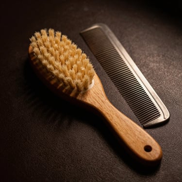 A wooden beard brush and a steel comb resting on a dark leather surface, warm side-lighting, masculine editorial style.