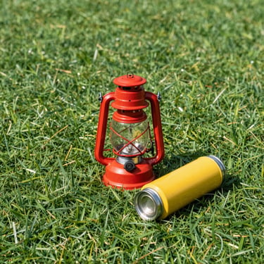 Bright outdoor flat-lay of a red camping lantern and a yellow steel thermos on clean green turf, crisp studio-style sunlight