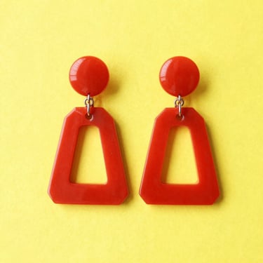 A pair of oversized geometric resin earrings, bright cherry red, isolated on a solid sunny yellow backdrop, high-key studio lighting