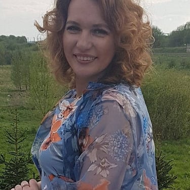 Smiling woman with curly red hair wearing a blue floral blouse outdoors in nature.