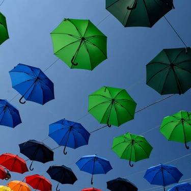 A photograph featuring numerous multicoloured umbrellas displayed in a rainbow pattern, floating serenely against a blue sky.