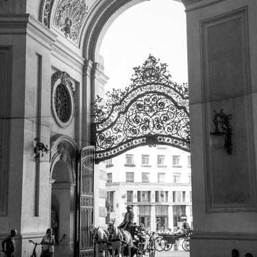 A black & white photo of people with bicycles and a man riding a carriage with horses passing under an arch.