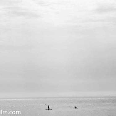 A black & white photograph of two paddle boarders gliding through tranquil sea waters at sunset under a dramatic sky