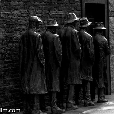 A black & white photo of five life-size men statues made of bronze, wearing hats and waistcoats, waiting in a queue by a wall
