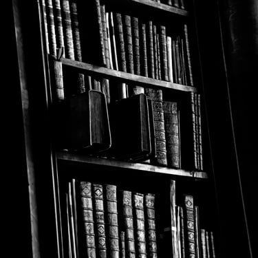 A black & white photo of an array of old library books meticulously arranged on a rustic wooden bookshelf