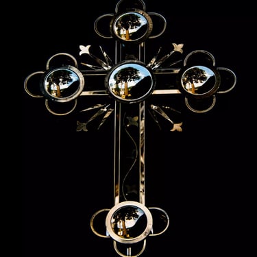A colour photograph of an exquisitely crafted cross made of reflective metal, mirroring the tree landscape opposite of it.
