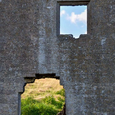 A colour photograph of a Medieval wall with a window at the bottom showing a patch of grass and at the top showing a blue sky