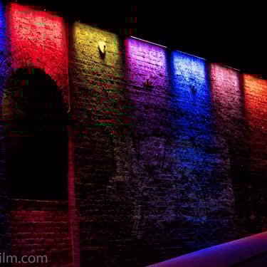 An urban night time photo of an old brick wall lit by an array of bold, bright lights in a spectrum of vivid rainbow hues