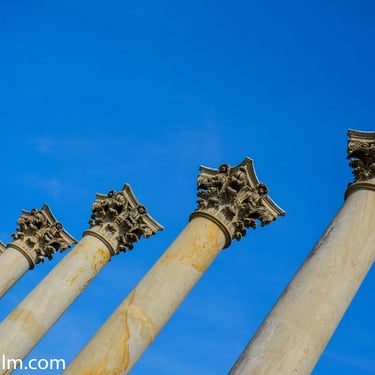 A colour photograph taken from below featuring six Greek style pillars set against a clear blue sky