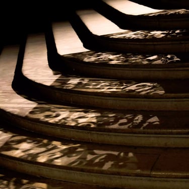 A sepia toned photograph of a dimly lit grand staircase with its masterfully crafted banister casting a thick shadow over it.