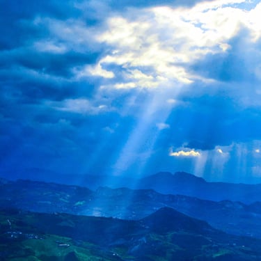 A colour photograph of a dramatic, lush green mountain landscape with sun rays breaking through thick and moody clouds