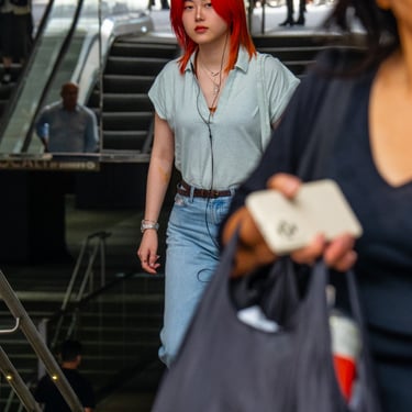 A woman with red hair walks up a stairway, behind another person. Escalators in the background create a busy setting.