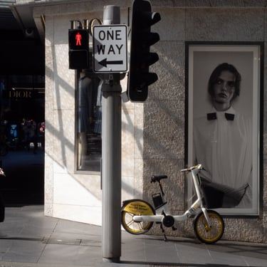 A street corner with a traffic light. A rental bike is parked, beside a chic fashion poster. Sunlight casts shadows.