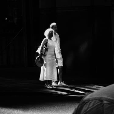 An elderly couple in light clothing, walking hand in hand on a sunlit street. They convey a sense of calm and companionship.