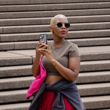 A person with short blonde hair wears sunglasses and a taupe top, holding a phone. She stands in front of wide stone steps.