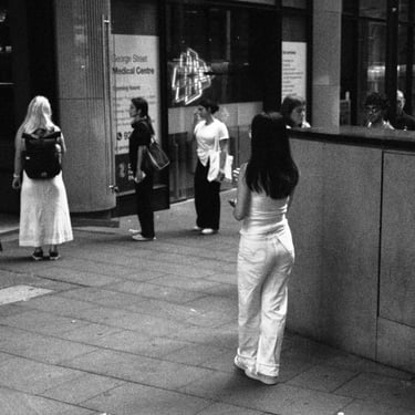A street scene with several people standing. A woman leans against a wall, while others converse nearby. Urban, candid.