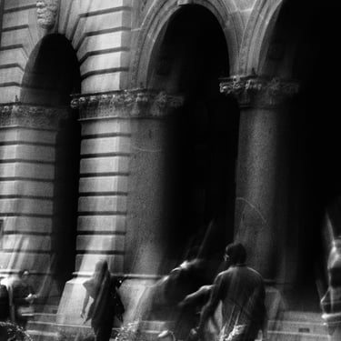 Black and white photo of people moving in front of a grand, arched, stone building facade, conveying motion and timelessness.