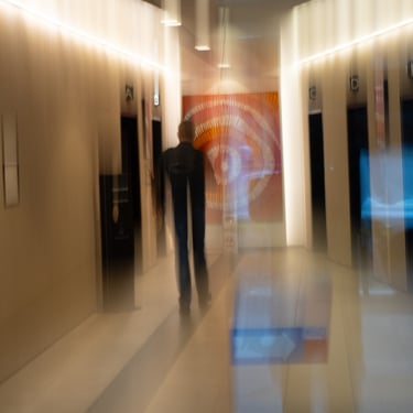 Image of a person walking down a hallway with elevator doors. The scene has a dreamy, abstract feel with soft lighting.