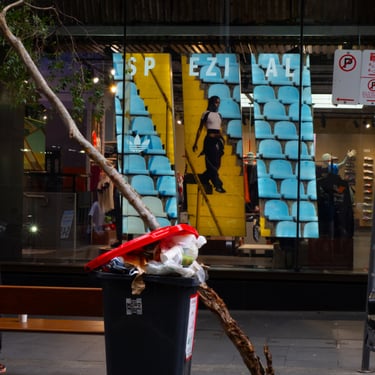 A trash bin overflowing with garbage and a fallen branch in front of a store window displaying vibrant yellow and blue ads.