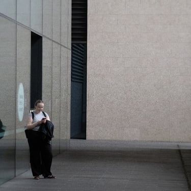 A person stands next to reflective glass buildings. The setting is minimalistic and the ambient tone is contemplative.