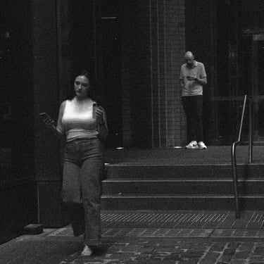 Photo of a woman walking with a smartphone in hand, while a man stands atop steps, absorbed in his phone.